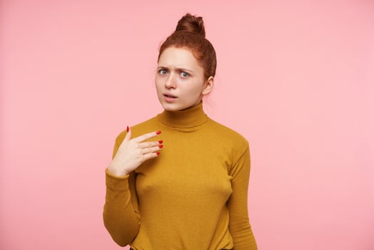 Indoor photo of young bewildered redhead female raising hand while looking confusedly at camera and frowning her eyebrows, standing over pink background