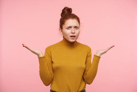 Puzzled young green-eyed redhead lady with natural makeup frowning her eyebrows and raising confusedly palms while looking at camera, standing over pink background photo
