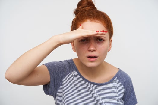 Studio photo of young redhead lady with natural makeup squinting her green eyes and raising hand to forehead while trying to see something far, isolated over pink background