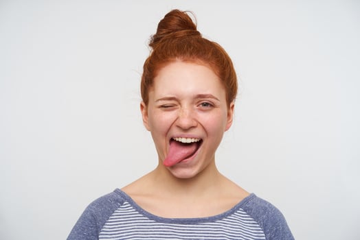 Joyful young loely redhead woman with natural makeup winking at camera and sticking happily out her tongue, having fun while posing over pink background photo