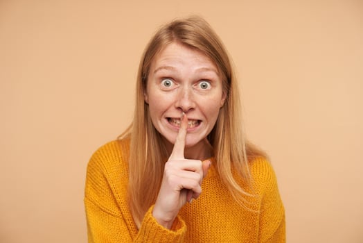 Indoor photo of young amazed redhead lady rounding her green eyes and holding raised forefinger on mouth while asking to keep secret, isolated over beige background