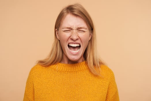 Stressed young long haired redhead female with casual hairstyle keeping her eyes closed while grimacing crossly her face, isolated over beige background photo