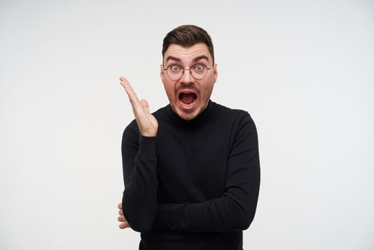 Bemused young dark haired bearded guy in glasses keeping his palm raised while looking excitedly at camera with wide mouth opened, isolated over white background photo