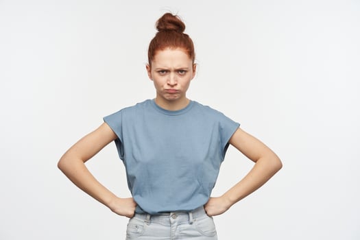 Portrait of upset, adult redhead girl with hair gathered in a bun. Wearing blue t-shirt and jeans. Put hands on her hips and frowning. Watching at the camera isolated over white background photo