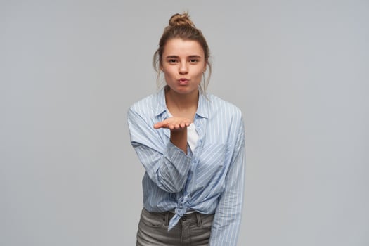 Teenage girl, pretty woman with blond hair gathered in bun. Wearing striped knotted shirt. Pursed her lips and sending air kiss over palm. Watching at the camera, isolated over grey background photo
