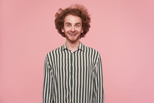 Young cool guy with brunette curly hair and beard. Wearing shirt with black and white stripes. Emotion and people concept. Excited, smile. Watching at the camera isolated over pastel pink background photo