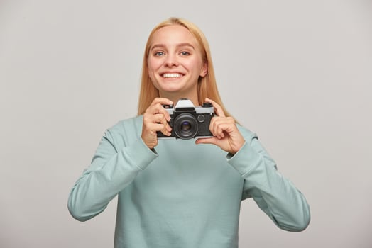 Young girl photographer looks happily smiling, holding a retro vintage photo camera in hands, in the process of shooting, working in studio, wearing blue casual sweatshirt, over grey background