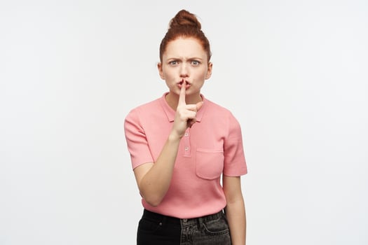 Angry looking woman, frowning redhead girl with hair bun. Wearing pink t-shirt and black jeans. Shows silence sign. Asking to be quiet. Watching at the camera, isolated over white background. photo