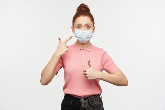Portrait of girl with ginger hair bun. Wearing pink t-shirt and medical, protective face mask. Pointing at the mask and shows thumb up. Watching at the camera, isolated over white background photo