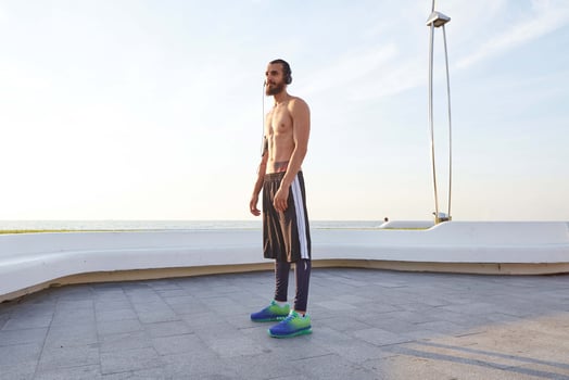 Young sporty bearded, rest after jogging, walks at the seaside, listen favorite song on headphones. Enjoys greates morning photo
