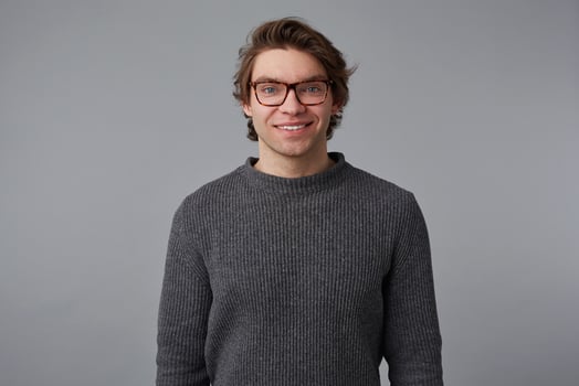 Photo of young cheerful handsome guy with glasses wears in gray sweater, stands over gray background and broadly smiles, looks happy and glad.