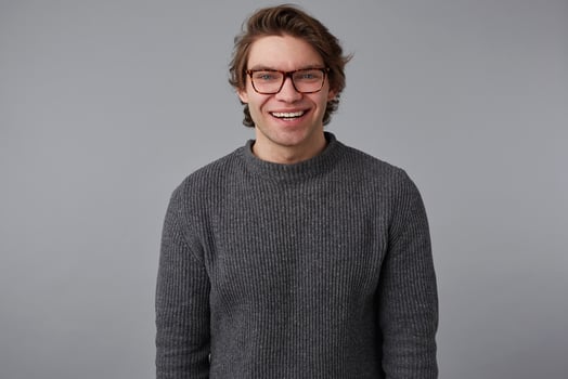 Photo of young positive handsome man with glasses wears in gray sweater, stands over gray background and broadly smiles, looks happy and cheerful.