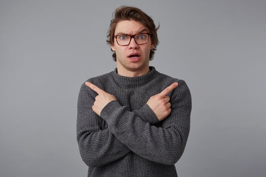 Portrait of young confused wondered man with glasses wears in gray sweater, stands over gray background, shows fingers in different directions. photo