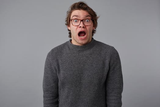 Portrait of young shocked guy with glasses, stands over gray background with wide open mouth and eyes, with surprised expression, looks scared. photo
