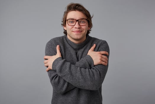 Portrait of young smiling man with glasses wears in gray sweater, stands over gray background and hugs himself, looks happines. photo