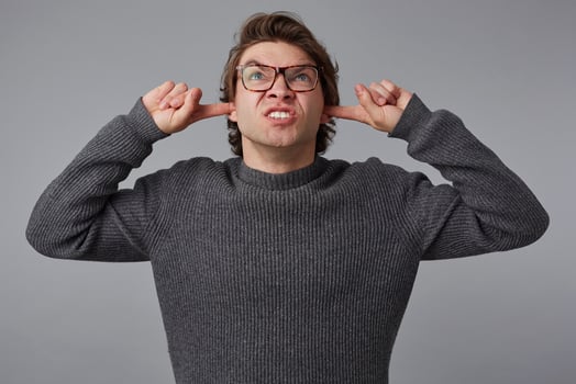 Portrait of young angry man with glasses wears in gray sweater, stands over gray background, disturbed by noise and covers ears with fingers. photo