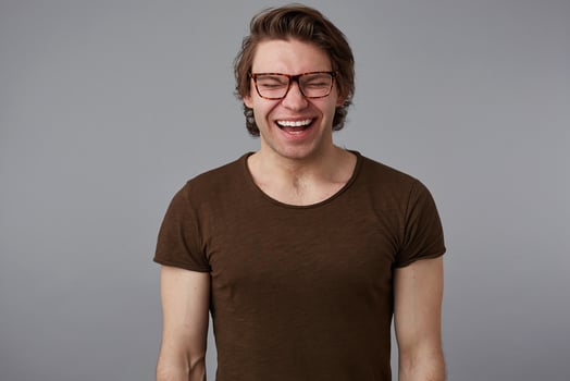 Photo of young loughting handsome man with glasses wears in basic t-shirt, stands over gray background and broadly smiles, looks happy and wondered.