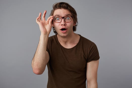 Portrait of young wondered man with glasses wears in basic t-shirt, stands over gray background and looks at the camera trough glasses. photo