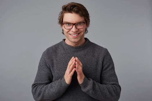 Young guy with glasses wears in gray sweater, stands over gray background, it should be folded palm and insidiously looking into the camera. photo