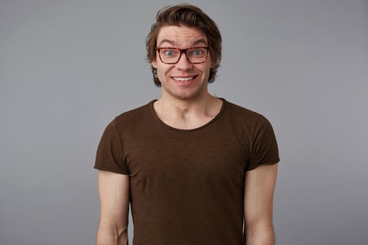 Photo of young surprised handsome man with glasses wears in basic t-shirts, stands over gray background and broadly smiles, looks happy and wondered.