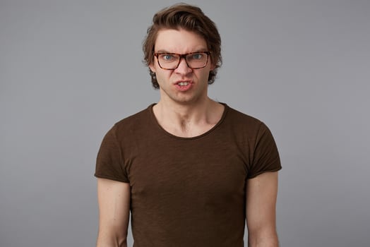 Photo of young displeased guy with glasses wears in blank t-shirt, stands over gray background and angry looks at the camera.
