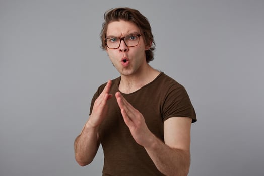 Photo of young guy with glasses wears in blank t-shirt standing in defensive posture, ready to punch, stands over gray background and try look expressing anger and fury.