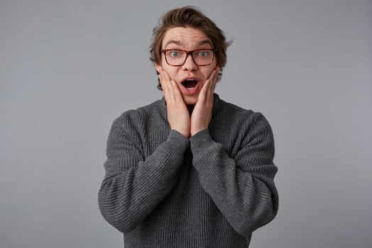 Young shocked guy with glasses wears in gray sweater, stands over gray background and touches cheeks with wide open mouth and eyes, hears amazed news, with surprised expression. photo