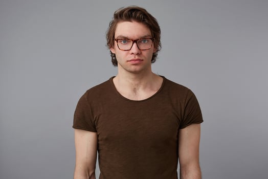 Photo of young serious guy with glasses wears in blank t-shirt, stands over gray background and looks at the camera without emotions.