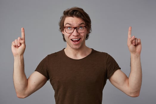 Portrait of young happy amazed guy with glasses, stands over gray background with surprised expression, points fingers up at a copy space over his head, looks at the camera and broadly smiles. photo
