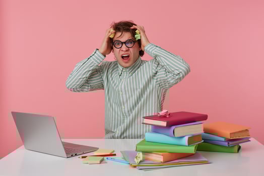 Studio shot of young crazy man guy with glasses, wears on blank shirt, sitting at a table with books, working at a laptop, looks angry and dissatisfied, something wrong. Isolated over pink background. photo