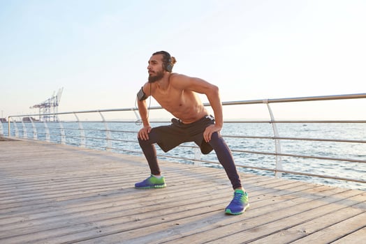 Young masculine sporty bearded guy doing stretching, morning exercises by the sea, listening favorite music on headphones, warm-up after run, leads healthy active lifestyle. Fitness male model. photo