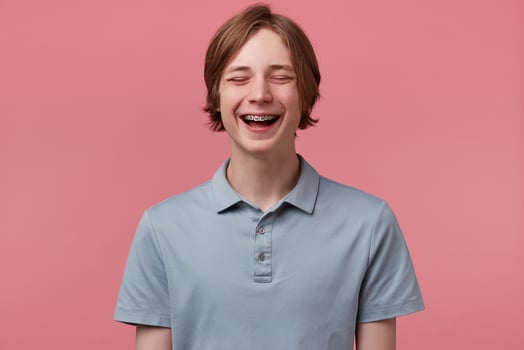 Close up of nice blue-eyed neatly combed young guy with braces on teeth joyfully laughing closed his eyes of fun wears polo t-shirt looks happy isolated over pink background photo