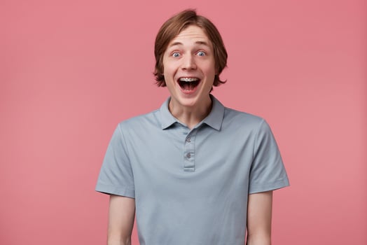 Close up of amazed excited young man with long neatly combed hair and braces on teeth wears polo t-shirt shouting and feels happy surprised isolated over pink background photo