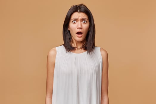 Portrait of scared woman in panic nervous frightened dressed in white dress, opened mouth feals fear, isolated on beige background photo