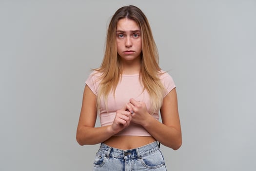 Unhappy girl, disappointed woman with blond long hair. Wearing pink t-shirt and jeans. People and emotion concept. Nervously touching fingers. Watching at the camera isolated over grey background photo