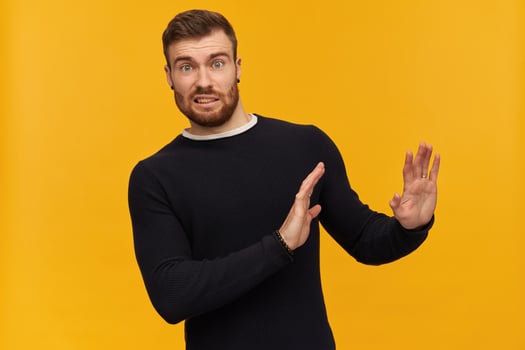 Bearded shocked guy, skeptical looking man with brunette hair. Has piercing. Wearing black sweater. Pushes arms away, reject gesture. Watching at the camera, isolated over yellow background photo