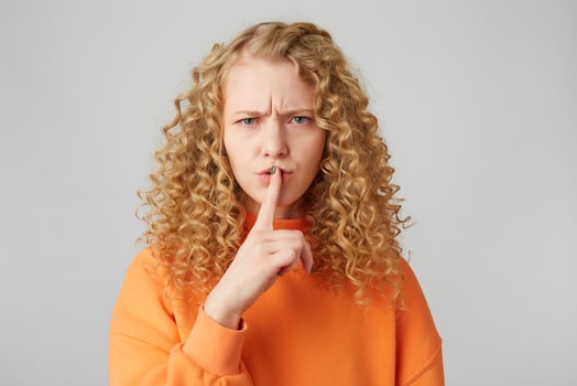 Serious female with curly blonde hair, keeps fore finger on lips, tries to keep conspiracy, looks camera, says Shh, make silence please. Isolated shot of girl shows silence gesture photo
