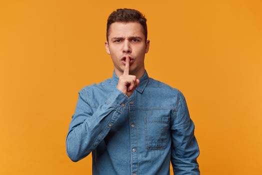 Seriously looking guy in the denim shirt urges everyone to keep silence, stay calm, presses his index finger to his lips depicts a gesture of silence, over yellow background photo