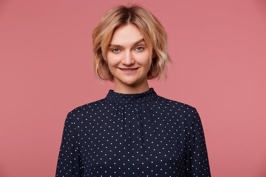 Close up of playful young beautiful attractive blonde woman dressed in blouse with polka dots, coquets, flirting, biting lip shows attraction, isolated over pink background photo