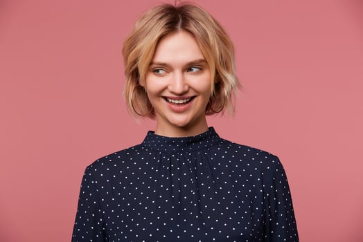 Nice young beautiful attractive blonde woman dressed in blouse with polka dots, has glad face expression, smiling, talking coquettishly looking to the side, isolated over pink background photo