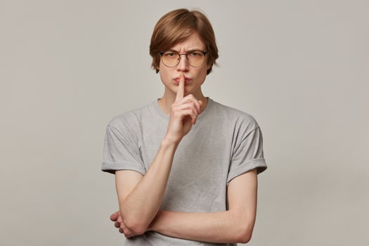 Portrait of handsome, angry male with blond hair. Wearing grey t-shirt and glasses. People and emotion concept. Showing silence sign and frowns. Watching at the camera isolated over grey background photo