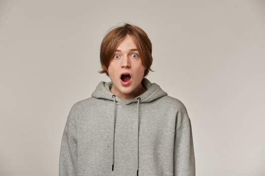 Portrait of shocked, amazed male with blond hair. Wearing grey hoodie. People and emotion concept. Stunned from what he sees. Watching surprised at the camera isolated over grey background photo