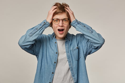 Portrait of stressed, adult male with blond hair. Wearing denim shirt and glasses, has braces. Holding hands on his head, frustrated. Watching at the camera isolated over grey background photo