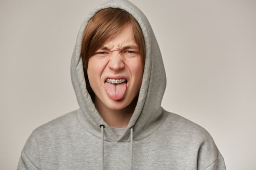 Portrait of displeased, adult male with blond hair. Wearing grey hoodie and has braces. Puts hood on and showing tongue. Emotion concept. Watching frowning at the camera isolated over grey background photo