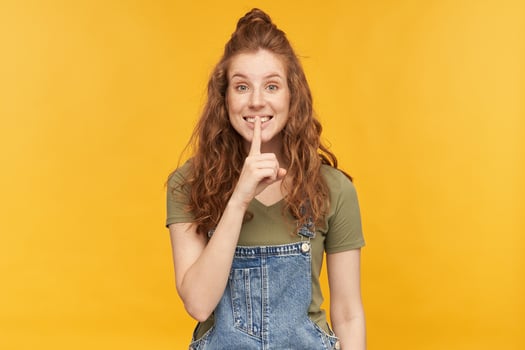Indoor portrait of young ginger female wears blue denim overalls and green t-shirt smiles broadly, flirt with somebody and show silence gesture. Isolated over yellow background photo