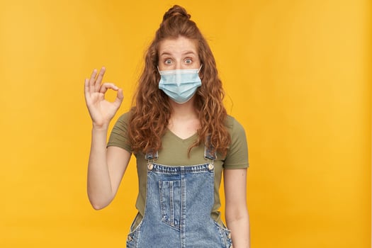 indoor studio shot of young ginger female with long red curly hair shows ok sign with positive, happy facial expression, wears face mask, isolated over yellow background photo