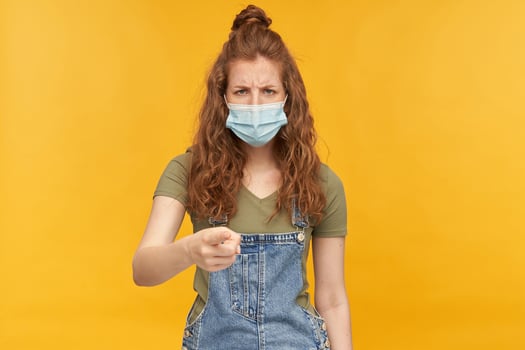indoor studio shot of young ginger female with long red curly hair points with a finger into camera with angry facial expression, wears face mask isolated over yellow background photo