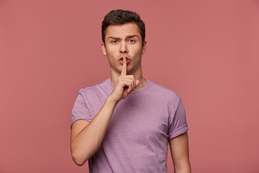 Phortrait of young attractive guy in blank t-shirt, looks at the camera, stands over pink background and shows silence gesture. photo