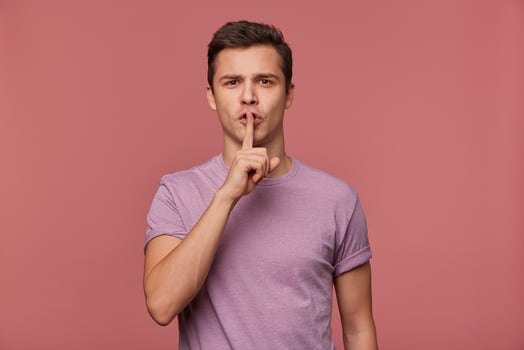 Photo of young attractive man in blank t-shirt, looks at the camera, stands over pink background and shows silence gesture.