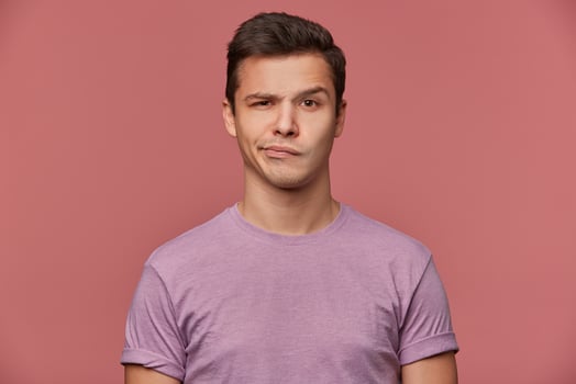 Portrait of handsome frowning young man wears in blank t-shirt, looks at the camera with with a grin and doubts, stands over pink background. photo